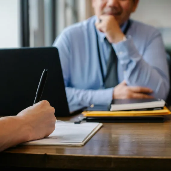 An interviewer takes notes on a clipboard across the desk from a candidate